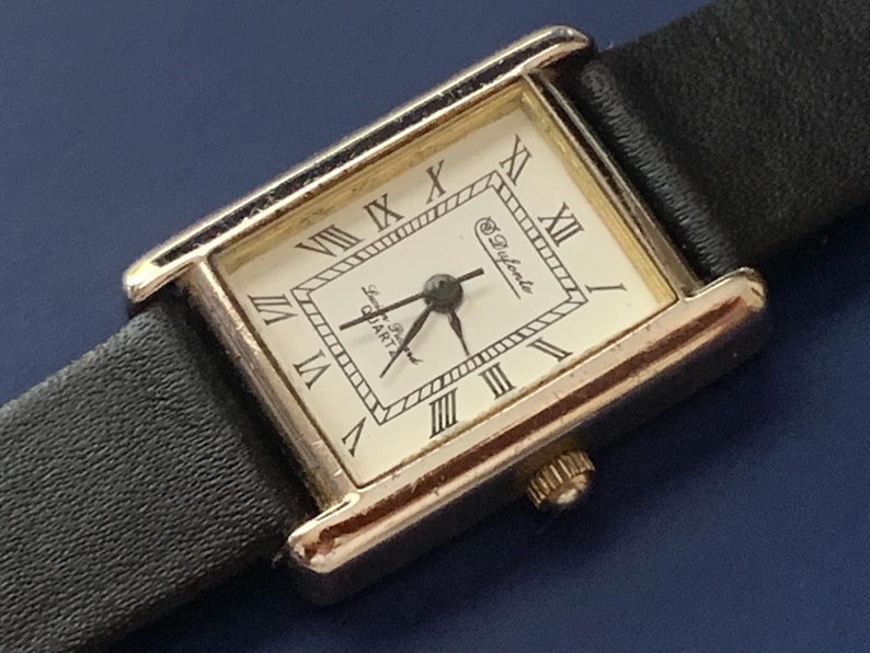 May include: A gold-tone rectangular watch with a white face and black leather band. The watch face has Roman numeral hour markers and the text "Le D'yante Quartz" printed on it.