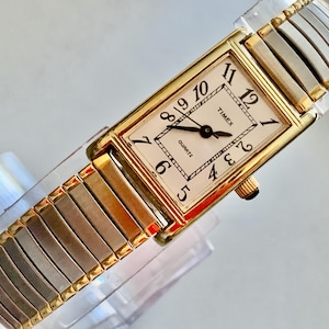 May include: A rectangular gold-toned Timex watch with a cream-colored face and black numerals. The watch band is a combination of gold and silver-toned metal. The watch face displays the word "TIMEX" and "QUARTZ".