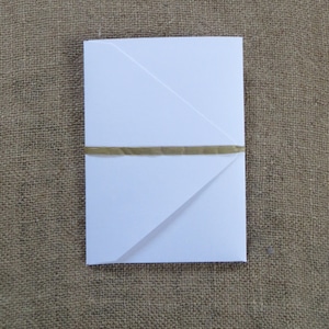 Pode incluir: Um envelope branco com uma fita dourada amarrada ao redor. O envelope est&aacute; dobrado em forma triangular.