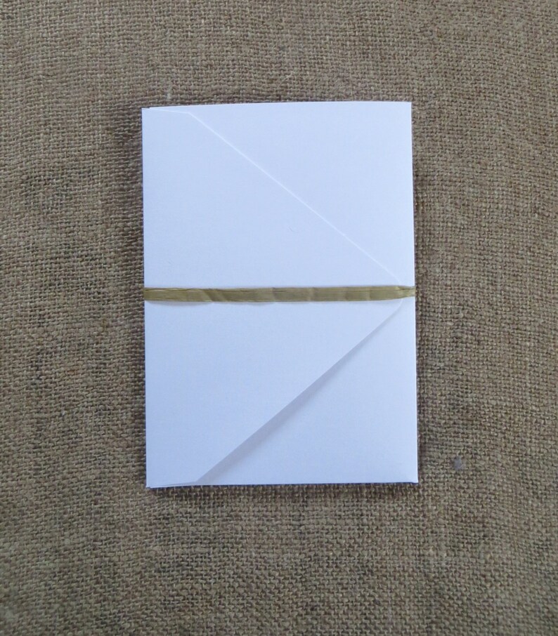 Pode incluir: Um envelope branco com uma fita dourada amarrada ao redor. O envelope est&aacute; dobrado em forma triangular.