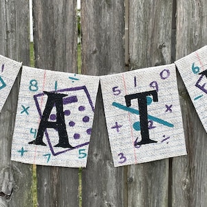 May include: A burlap banner with the word "MATH" spelled out in black letters. Each letter is on a separate flag with a white background and various math symbols and numbers printed on it.