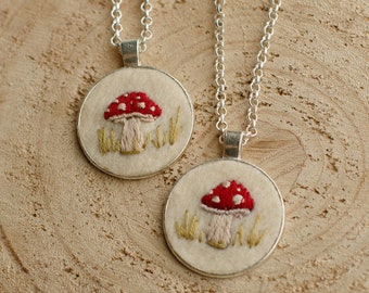 May include: Two silver necklaces with round pendants featuring embroidered red and white mushrooms on a white background. The mushrooms are surrounded by green embroidery that resembles grass.