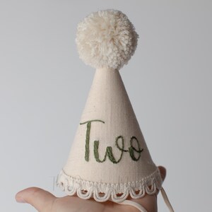 May include: A cream-colored birthday party hat with a fluffy pom-pom on top. The hat is cone-shaped with the word "Two" embroidered in green script. The bottom edge has a scalloped lace trim.