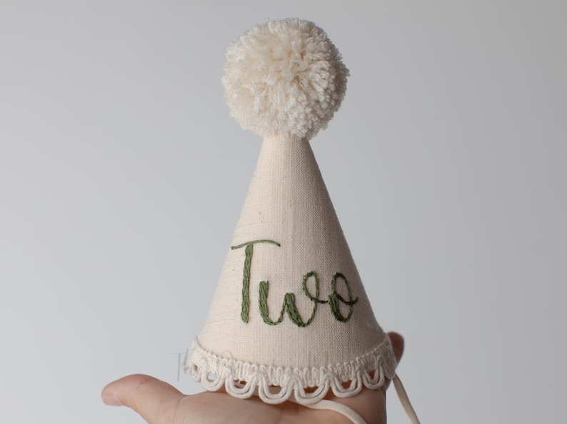 May include: A cream-colored birthday party hat with a fluffy pom-pom on top. The hat is cone-shaped with the word "Two" embroidered in green script. The bottom edge has a scalloped lace trim.