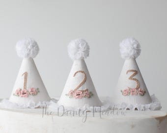 May include: Three white linen party hats with pink flowers and burlap numbers 1, 2, and 3. The hats have white pom poms on top.