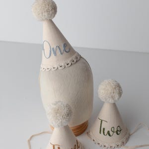 May include: Off-white party hats with pom-poms and embroidered numbers: One, Two, and Three. The hats have a scalloped lace trim and are designed for a birthday celebration. The hats are made of a soft, light-colored fabric.