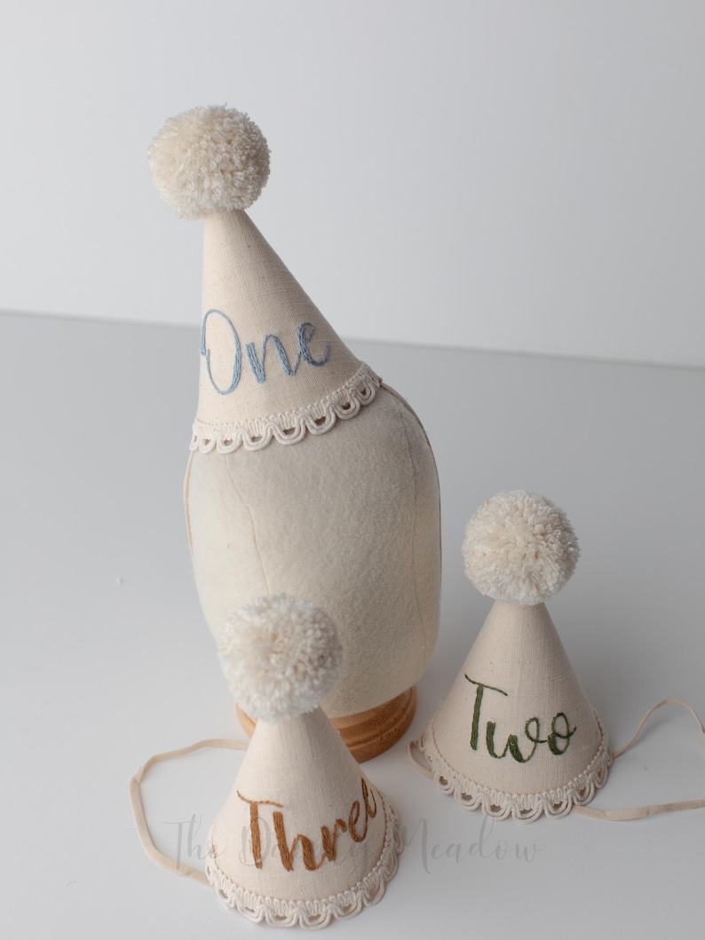 May include: Off-white party hats with pom-poms and embroidered numbers: One, Two, and Three. The hats have a scalloped lace trim and are designed for a birthday celebration. The hats are made of a soft, light-colored fabric.