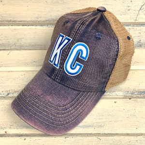 May include: A black and brown trucker hat with a blue and white embroidered "KC" on the front.