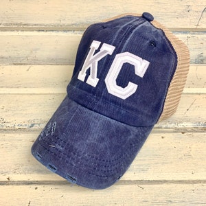 May include: A navy blue and tan distressed baseball cap with a white "KC" embroidered on the front.