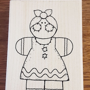 May include: A wooden stamp featuring a whimsical, hand-drawn illustration of a doll-like figure. The black outline depicts a doll with a bow, dress, and stitched details. The stamp is rectangular and made of light-colored wood.