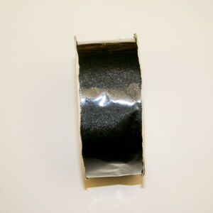 May include: A roll of black, textured tape with a shiny, metallic appearance. The tape is housed in a cardboard roll, and the image is taken from a top-down perspective.