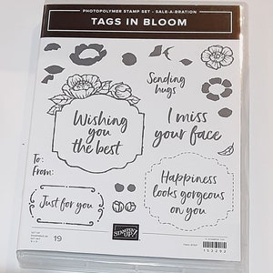 May include: A set of 19 photopolymer stamps featuring floral designs and sentiments. The stamps are packaged in a clear plastic case with the title "Tags in Bloom" and the phrase "Sale-A-Bration" printed on the front. The stamps include phrases such as "Wishing you the best", "Sending hugs", "I miss your face", and "Happiness looks gorgeous on you".