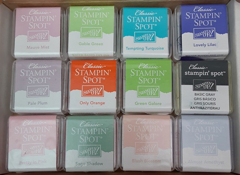 May include: Twelve small square ink pads in a clear plastic case. The ink pads are labeled with the brand name "Stampin' Spot" and various color names, including Mauve Mist, Gable Green, Tempting Turquoise, Lovely Lilac, Pale Plum, Only Orange, Green Galore, Basic Gray, Pretty in Pink, Sage Shadow, Blush Blossom, and Almost Amethyst.