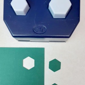 May include: A blue hexagon paper punch with two light gray hexagon-shaped push buttons. The punch is next to a green sheet of paper with two white hexagon cutouts and two green hexagon shapes.