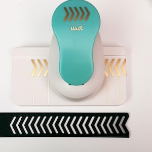 May include: A teal and white paper punch with gold arrow accents and the text "We R". A black paper strip with a cut-out arrow design is below the punch. The punch is on a white background.