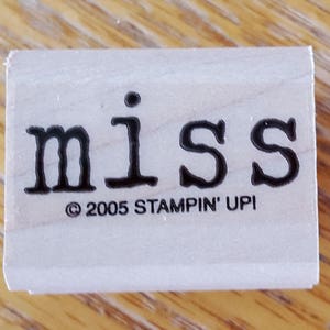 Könnte beinhalten: Ein rechteckiger Holzstempel mit dem Wort "miss" in schwarzer Serifen-Schrift. Darunter das Copyright-Zeichen, das Jahr "2005" und der Text "STAMPIN' UP!". Der Stempel befindet sich auf einer hellen Holzoberfläche.