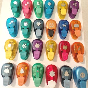 May include: A selection of colourful craft punches featuring diverse shapes such as a leaf, a coffee cup, a spider, a pumpkin, a cat, and a musical note. These plastic punches have a lever for ease of use.