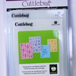 May include: A package of Cuttlebug embossing folders for use with a Cricut machine. The package includes four folders with different designs, including "Sew", "Rock", "Yum", and a floral design. The folders are designed to coordinate with the Cricut Country Life cartridge.
