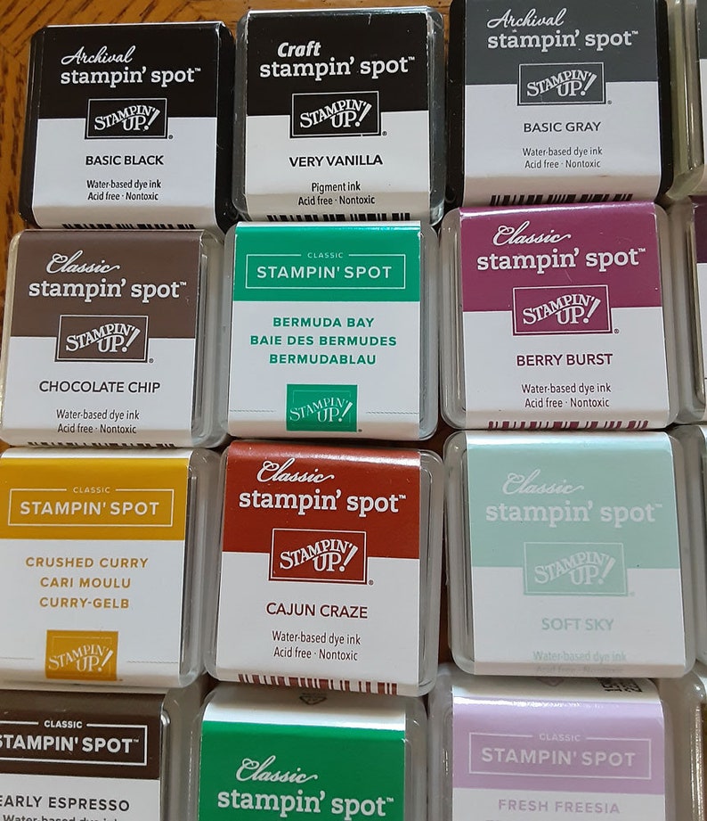 May include: A collection of 12 ink pads in various colors, including black, gray, brown, green, red, and purple. Each ink pad has a clear plastic case with a label that reads "stampin' spot" and "STAMPIN' UP!" in a white font. The ink pads are arranged in a grid pattern.