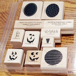 May include: A set of nine wooden stamps featuring Halloween-themed designs. The stamps include a pumpkin face, a swirl, a dotted circle, a striped circle, a pumpkin face with a tooth, a pumpkin face with eyes, a pumpkin face with a nose, a line of vertical lines, and the text "from our patch to yours - happy halloween!" and "sending halloween smiles across the miles!"