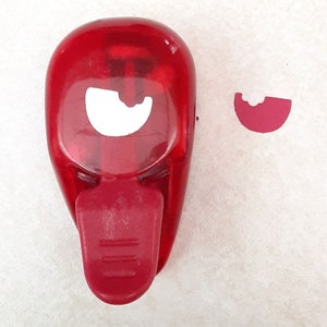 May include: A red plastic punch with a white crescent moon shape cut out. A red crescent moon shape has been punched out of a piece of paper.