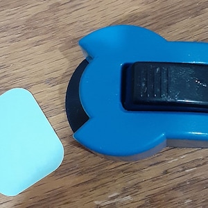 May include: A blue and black rotary cutter with a black blade and a light blue square adhesive pad. The cutter has a black sliding lock and is designed for cutting paper or fabric. The items are on a wooden surface.