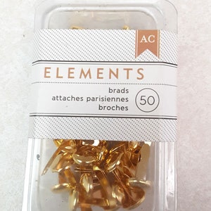 May include: A clear plastic package containing 50 gold-colored brads. The package has a white label with the word "ELEMENTS" and the text "brads, attaches parisiennes, broches". The brads are small, with a flat head and two prongs.