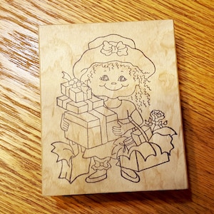 May include: A wooden stamp with a line drawing of a girl wearing a hat and carrying a stack of presents. The girl is smiling and has curly hair.