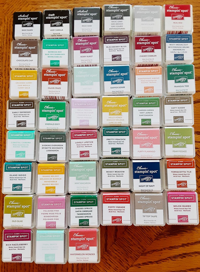 May include: A collection of 25 small, square, clear plastic containers with colorful ink pads inside. Each container has a label with the brand name "Stampin' Spot" and the ink color name. The ink pads are arranged in five rows of five containers.