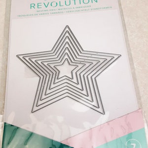 May include: A package of We R Memory Keepers Revolution nesting dies for crafting. The dies are silver and shaped like a star with seven points. The package is white with blue and pink accents. The text on the package reads "We R memory keepers. REVOLUTION™ NESTING DIES MATRICES A IMBRIQUER TROQUELES DE VARIOS TAMAÑOS VERSCHACHTELT STANZFORMEN 7 PC/PZ".
