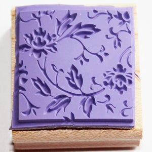 K&ouml;nnte beinhalten: Ein lilafarbener Gummistempel mit einem floralen Design. Der Stempel ist auf einem Holzblock montiert.