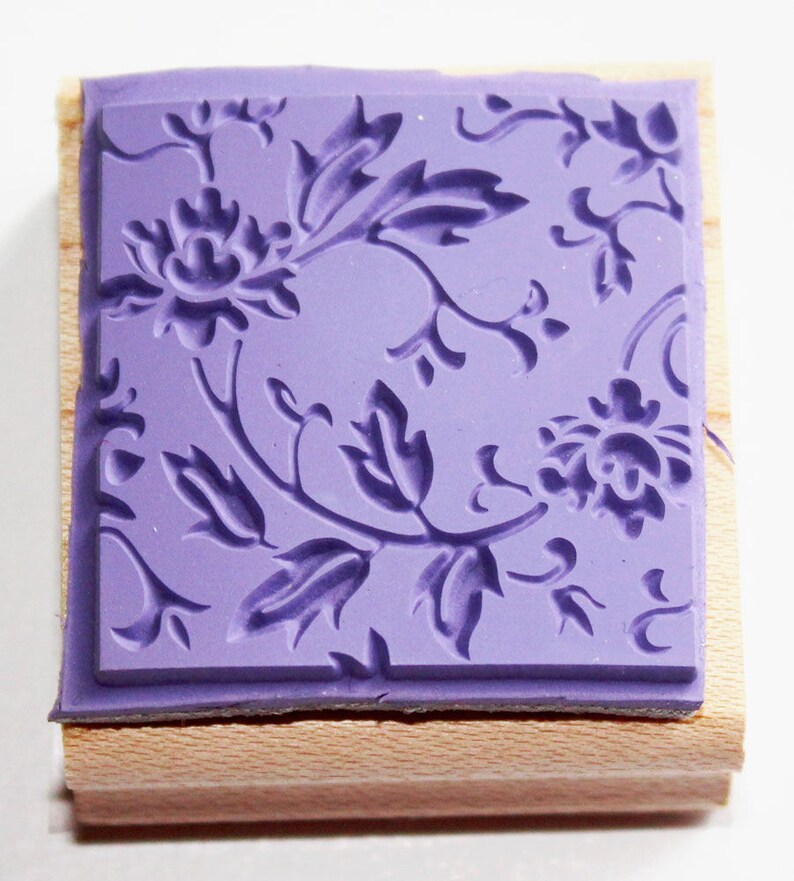 K&ouml;nnte beinhalten: Ein lilafarbener Gummistempel mit einem floralen Design. Der Stempel ist auf einem Holzblock montiert.
