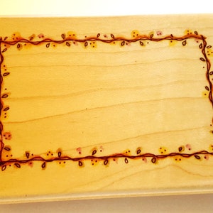 Puede incluir: Un sello de madera con un diseño de borde floral marrón. El borde está formado por una línea continua de enredaderas con pequeñas hojas y puntos.