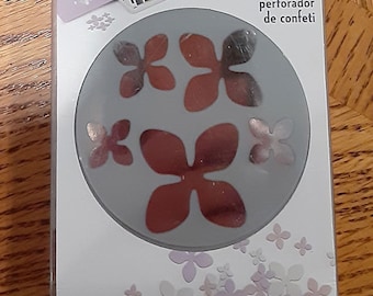 Peut inclure: Un gros plan sur un poinçon à confettis en forme de fleur avec un corps en plastique gris et un motif de flocon de neige argenté. Le poinçon est emballé dans une pochette en plastique transparente avec un fond jaune et le texte "confetti punch", "poinçons à confettis", et "perforador de confeti".