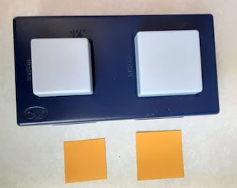 May include: A blue square paper punch with two light gray square buttons. The punch has the measurement "1 ½" printed on it. Two orange square paper cutouts are below the punch.