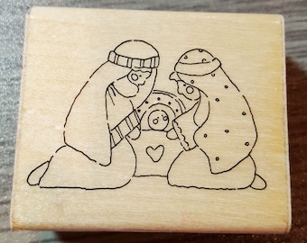 May include: A wooden rubber stamp with a black and white line drawing of the nativity scene. The image depicts Mary and Joseph kneeling beside a manger with a baby inside. A heart is drawn above the manger.