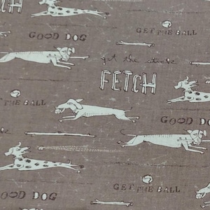Puede incluir: Tela marrón con un patrón repetido de perros corriendo, huesos y texto. El texto incluye "FETCH", "GOOD DOG" y "GET THE BALL". Los perros son blancos, algunos con manchas negras. El diseño tiene un aspecto vintage.