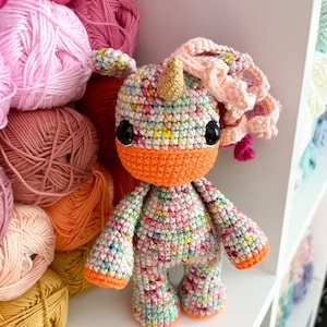 Könnte beinhalten: Ein farbenfrohes gehäkeltes Einhorn-Spielzeug mit einem goldenen Horn, orangefarbenen Akzenten und rosa Wollhaaren. Das Einhorn sitzt auf einem weißen Regal mit Wollknäueln im Hintergrund.