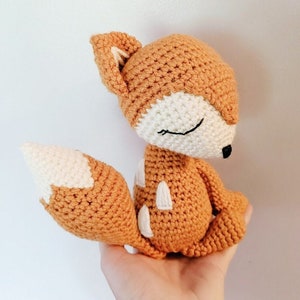 May include: A crocheted fox plush toy with a brown and white color scheme. The fox has closed eyes and a small, pointed nose.