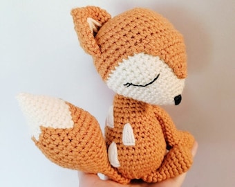 May include: A crocheted fox plush toy with a brown and white color scheme. The fox has closed eyes and a small, pointed nose.