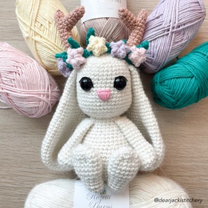 May include: White crocheted bunny with a floral crown and antlers. The bunny is sitting on a table with yarn balls in various colors.