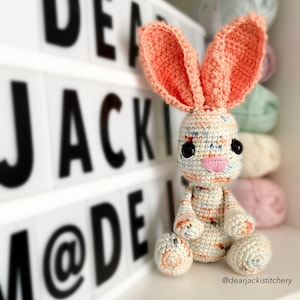 May include: A handmade crochet bunny with large, upright peach-colored ears and a speckled body. The bunny has black button eyes and a pink embroidered nose. The background includes a light box with the text "DEAR JACKIE MODE" and a shelf with yarn.