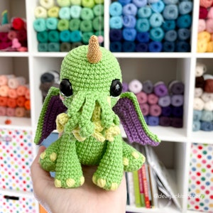 May include: A green crocheted Cthulhu plush toy with purple wings and yellow tentacles. The toy has large black eyes and a gold horn on its head. The toy is sitting on a hand.