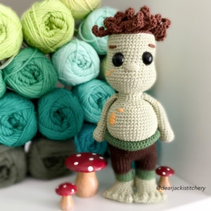 May include: A crocheted green and brown gnome doll with a brown curly hair, brown eyes, and a brown belt. The gnome is standing on a white surface with two red and white mushroom figurines.