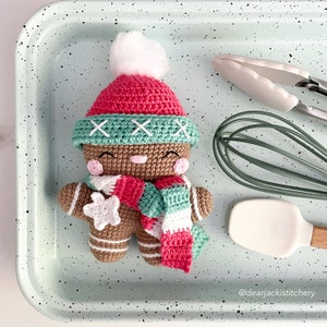 Pode incluir: Um boneco de gengibre de crochê vestindo uma echarpe listrada vermelha e branca, um chapéu verde com pontos brancos e um pompom branco. O boneco de gengibre está segurando uma estrela branca de crochê. O boneco de gengibre está sentado em uma superfície verde claro com manchas pretas. A imagem inclui o texto "@dearjackstitchery".