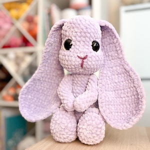 May include: A crocheted plush bunny toy with large floppy ears. The bunny is light purple with black eyes and a pink nose. The bunny is sitting on a wooden surface.