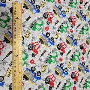 May include: Fabric with a repeating pattern of colorful construction vehicles, traffic lights, and road signs on a light gray background. The vehicles are red, blue, and green. The ruler shows inches.