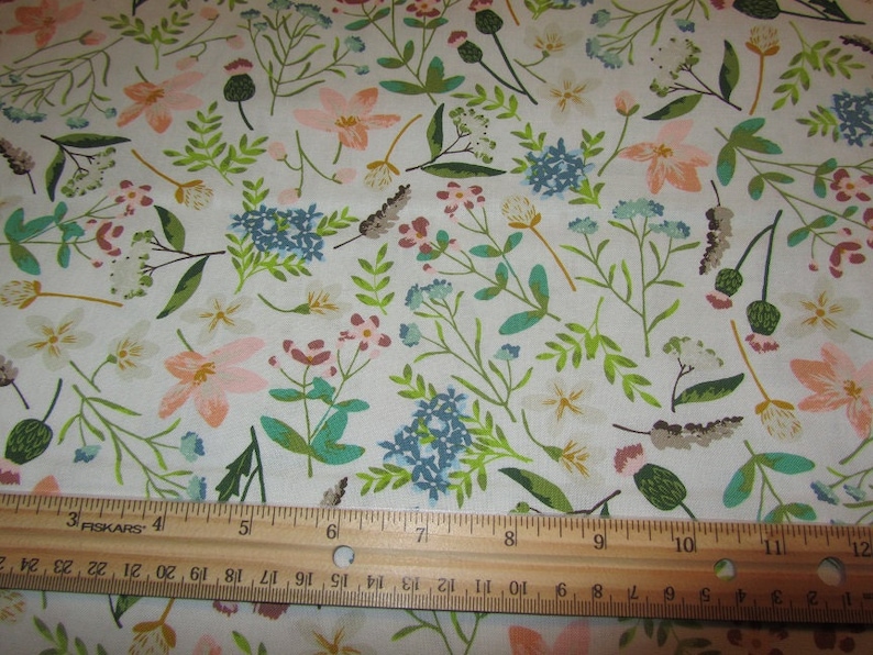 May include: White fabric with a repeating pattern of pink, blue, and green flowers. A wooden ruler with the text "FISKARS" is placed at the bottom of the image.