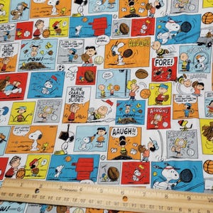 May include: White fabric featuring a colorful comic strip design with characters like Snoopy and Charlie Brown. The panels depict various sports scenes with text bubbles and action words like "AAUGH!" and "FORE!" A wooden ruler is placed on the fabric.