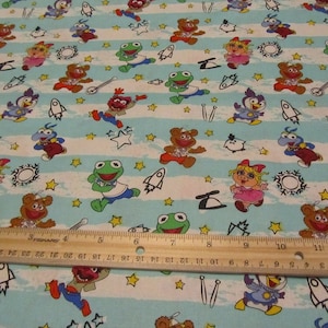 May include: Blue and white striped fabric with a repeating pattern of Muppet characters including Miss Piggy, Kermit the Frog, Animal, and Fozzie Bear. The characters are running and jumping with stars and rockets in the background.